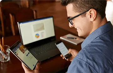 Laptop open on a desk with a man looking at a tablet whilst also holding a mobile phone.