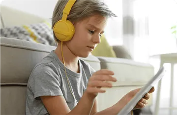 Young child wearing yellow heaphones leaning up against a sofa looking on a tablet.