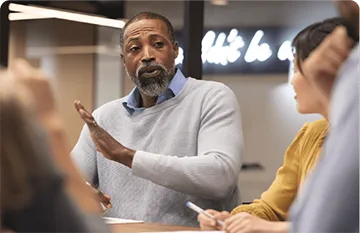 Man wearing a shirt and jumper sitting behind a desk talking to a grop of people.